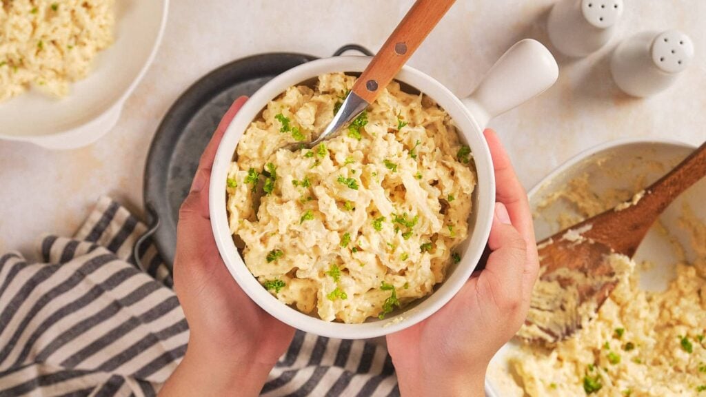 Hands holding a white bowl of cauliflower mac and cheese garnished with chopped herbs.