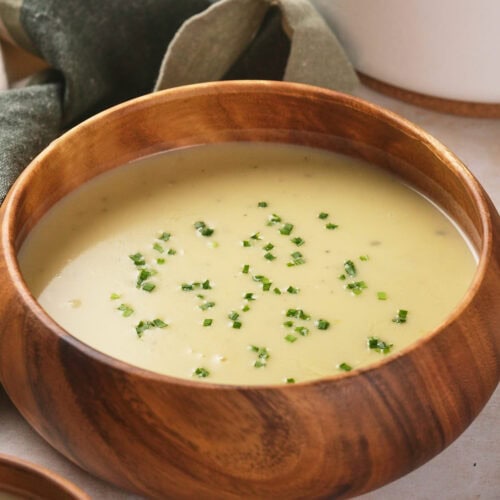 Wooden bowl of potato and leek soup garnished with chopped chives, placed on a light surface.