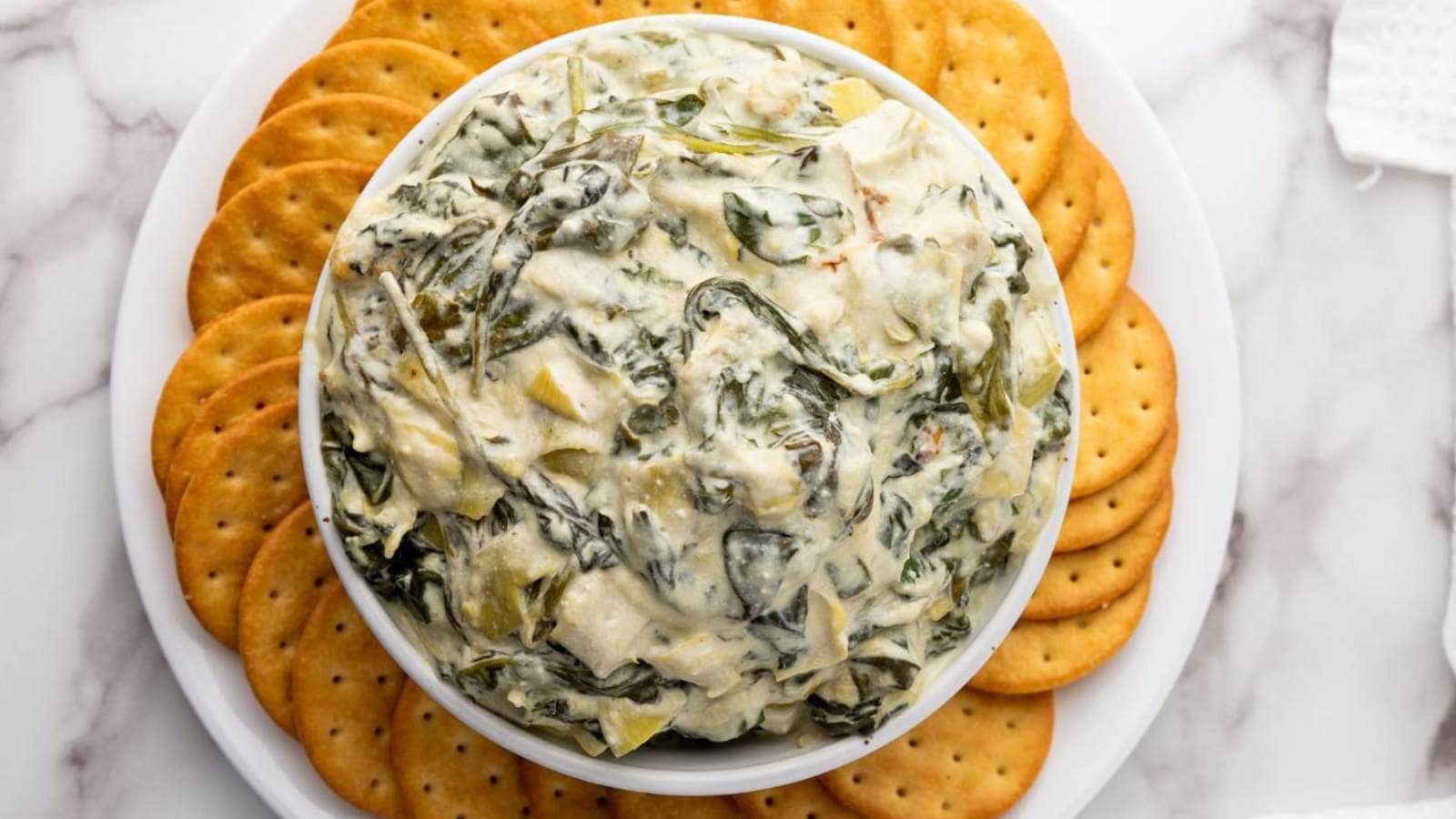 A bowl of spinach artichoke dip surrounded by round crackers on a white marble surface.