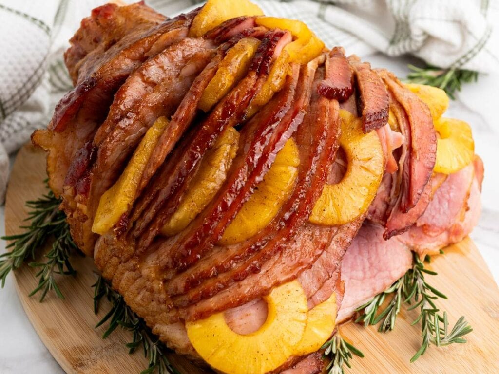Sliced ham topped with pineapple rings arranged on a wooden board, garnished with rosemary sprigs.