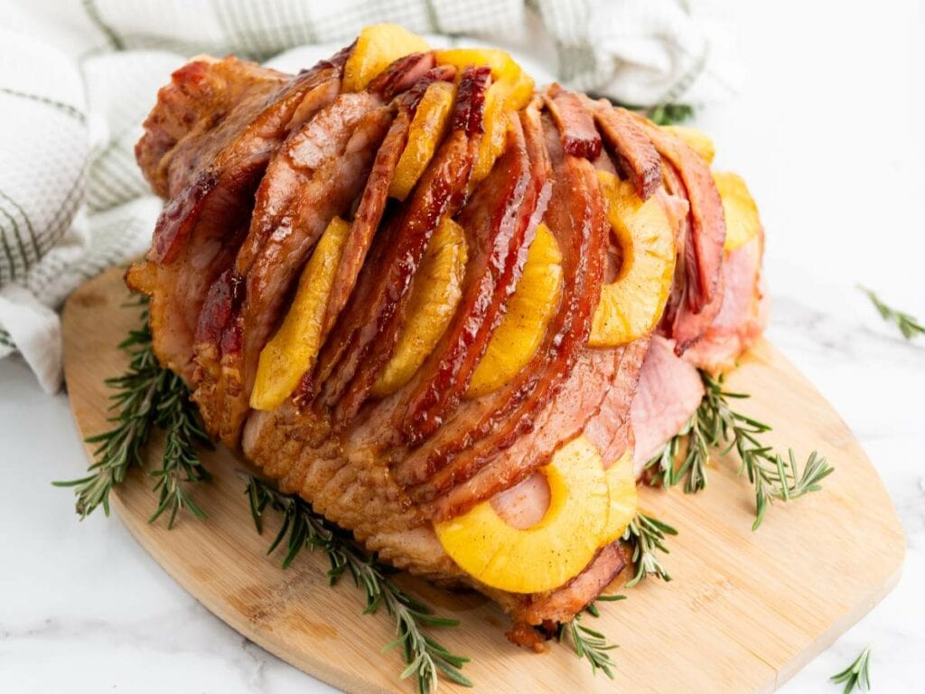 A sliced holiday ham garnished with pineapple and rosemary on a wooden board.