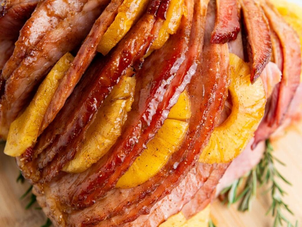 Close-up of a glazed holiday ham with slices of pineapple and rosemary on a light wooden surface.