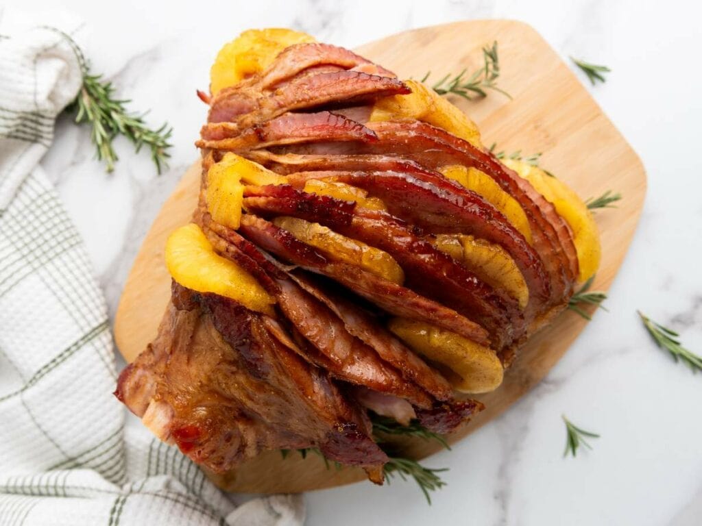 Sliced glazed holiday ham with pineapple rings on a wooden cutting board, garnished with rosemary sprigs, next to a checkered cloth.