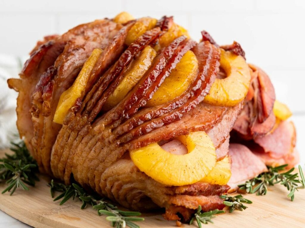 A glazed holiday ham topped with pineapple slices and garnished with rosemary, placed on a wooden board.