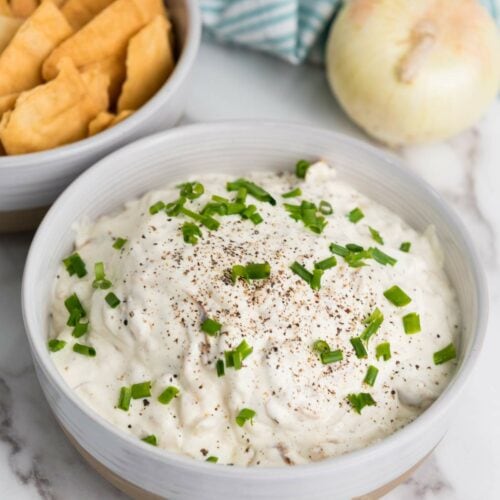 A bowl of creamy caramelized onion dip topped with chopped green onions and black pepper, next to a bowl of crackers. An onion and striped cloth are in the background.