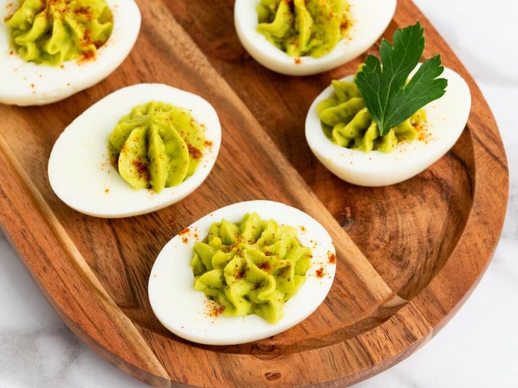 Deviled eggs topped with green filling and sprinkled with paprika, served on a wooden tray, with a parsley garnish on one egg.