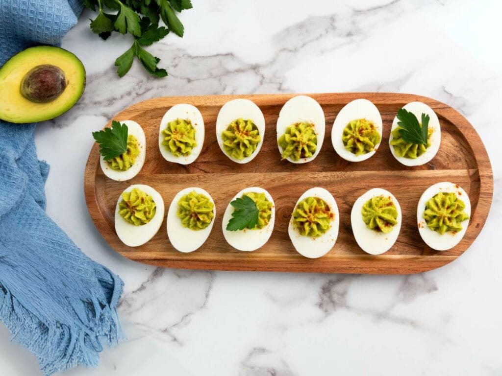 Wooden platter with twelve deviled eggs topped with avocado mixture and parsley leaves.