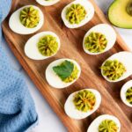 Deviled eggs on a wooden tray, topped with green filling and garnished with parsley, placed beside a blue cloth and half an avocado.