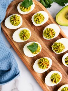 Deviled eggs on a wooden tray, topped with green filling and garnished with parsley, placed beside a blue cloth and half an avocado.
