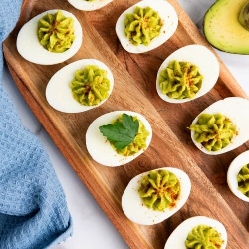 Deviled eggs on a wooden tray, topped with green filling and garnished with parsley, placed beside a blue cloth and half an avocado.