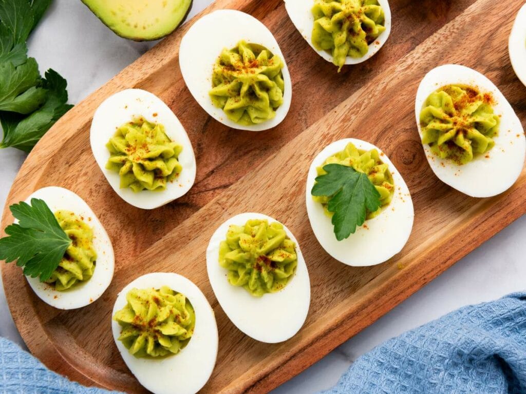 Deviled eggs with green filling and paprika garnish on a wooden platter, some topped with parsley leaves.