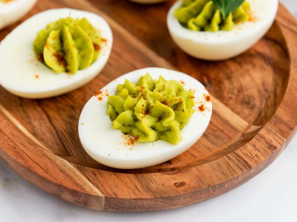 Deviled eggs with a green filling, garnished with paprika, arranged on a wooden plate.
