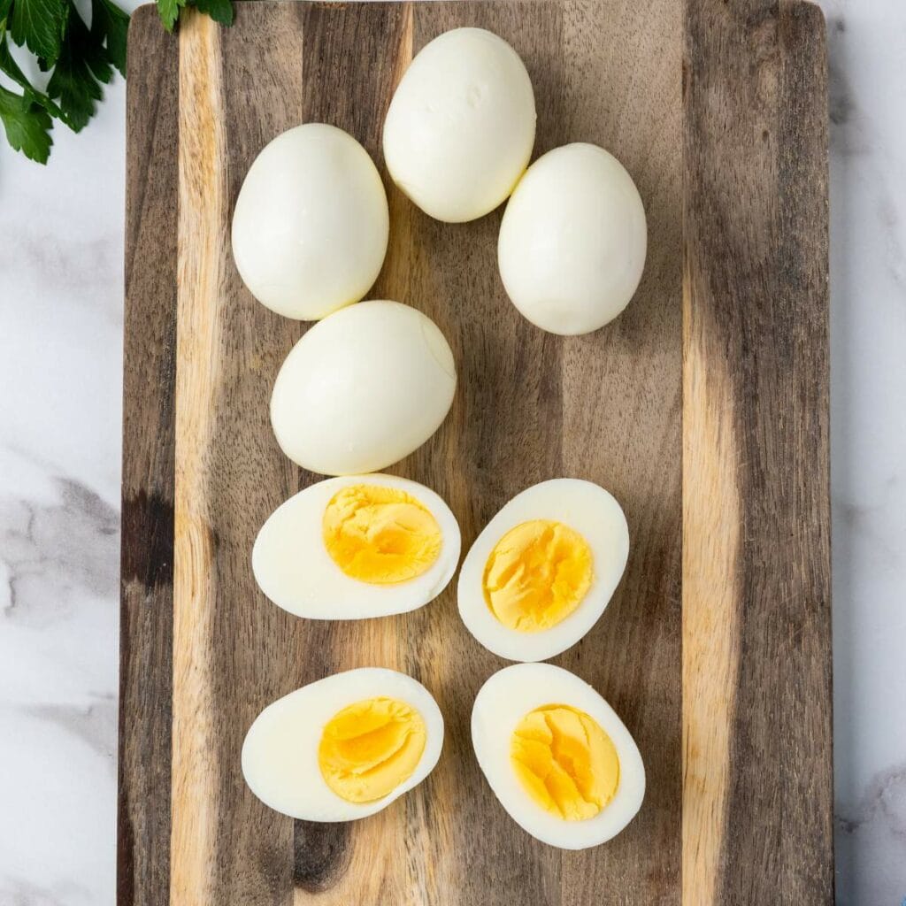 Seven hard-boiled eggs on a wooden cutting board, three are halved showing yolks. Parsley partially visible.