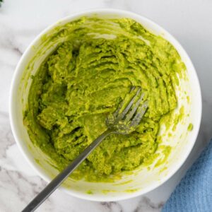 A bowl of mashed avocado with a fork, placed on a marble surface.