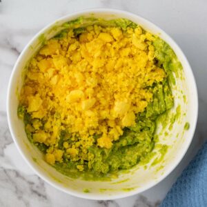 Bowl of mashed avocado topped with crumbled yellow egg yolk on a marble surface.