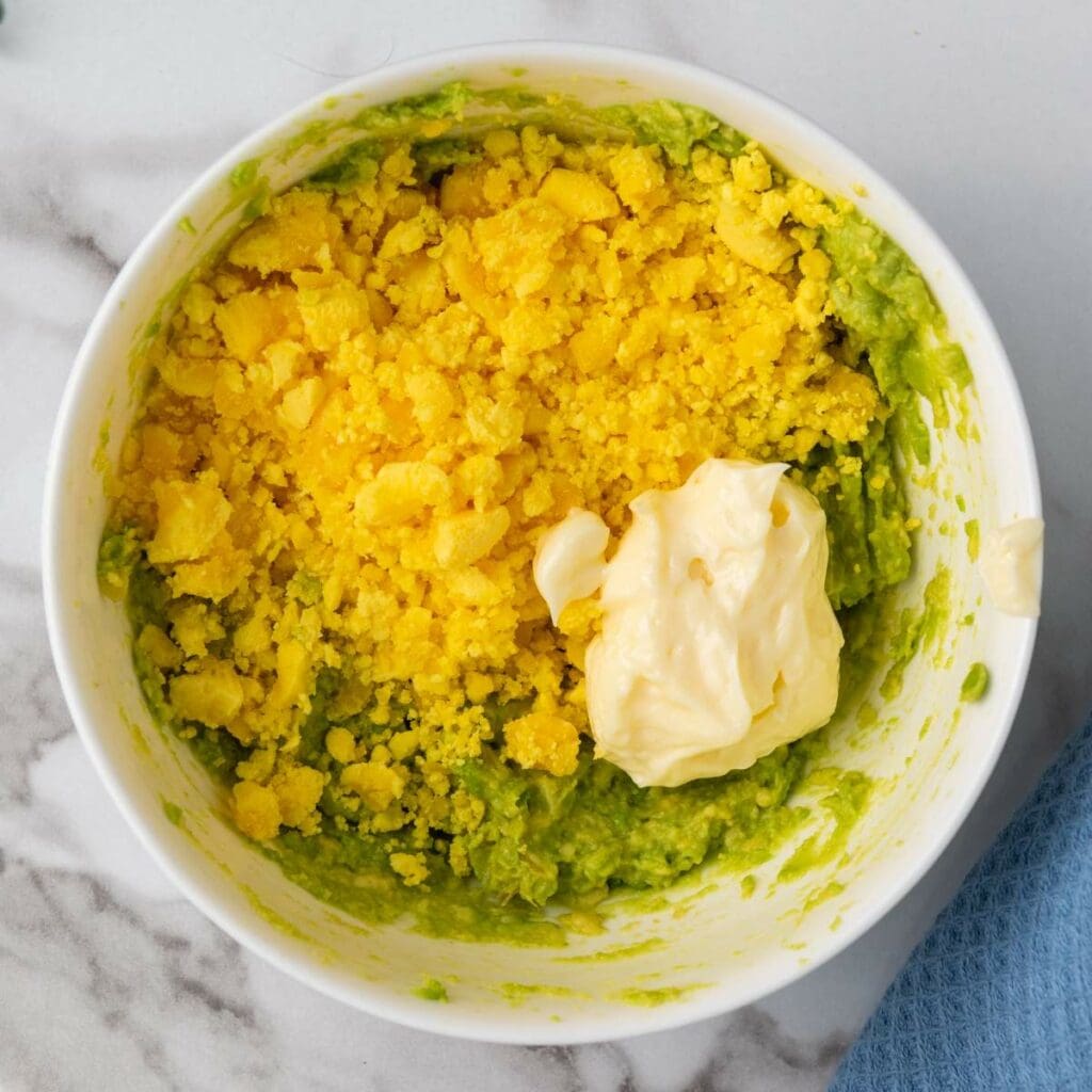 A bowl containing mashed avocado, crumbled egg yolks, and a dollop of mayonnaise.