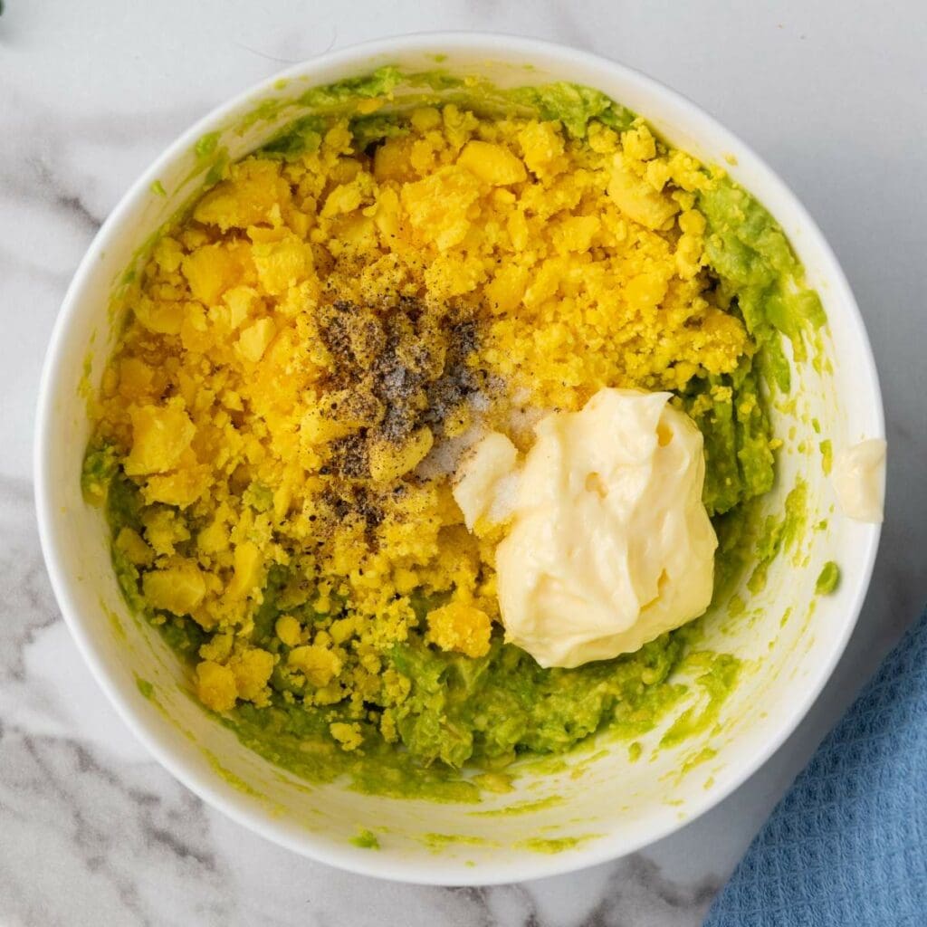 Bowl containing a mixture of mashed avocado, grated egg yolk, mayonnaise, salt, and black pepper on a marble surface.