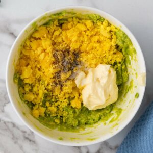 Bowl containing a mixture of mashed avocado, grated egg yolk, mayonnaise, salt, and black pepper on a marble surface.