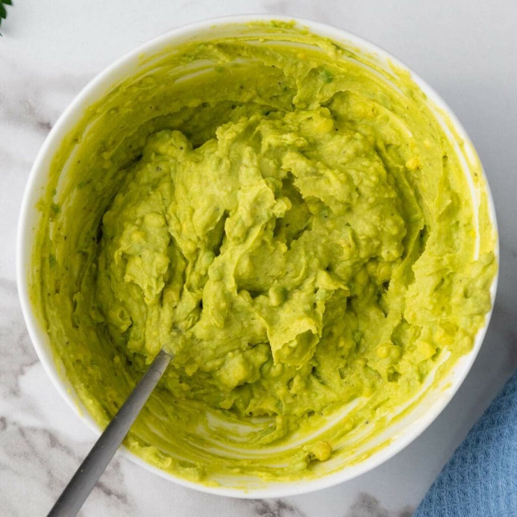 A bowl of guacamole with a spoon, placed on a marble surface.