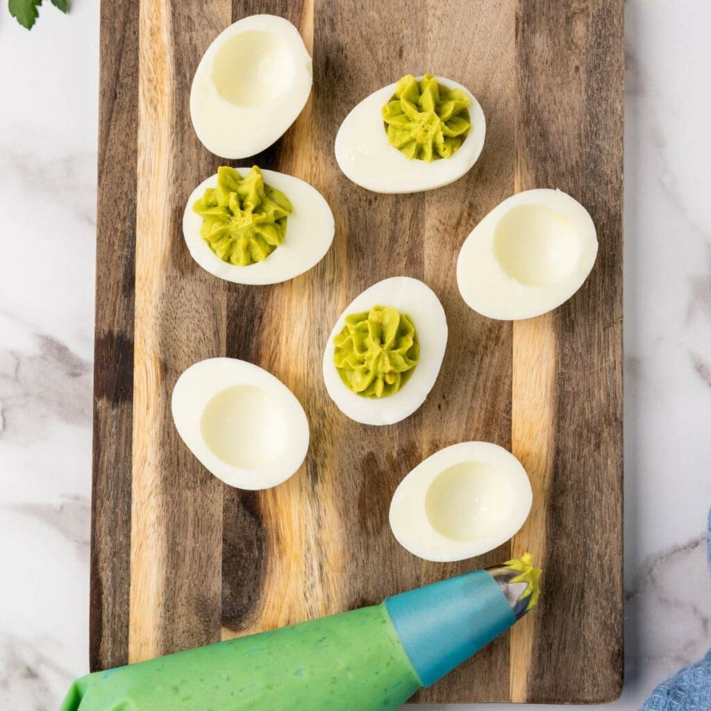 Hard-boiled egg halves on a wooden board, with some filled with green avocado mixture using a piping bag.