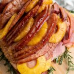 A glazed holiday ham adorned with pineapple slices sits on a wooden board, garnished with sprigs of rosemary.