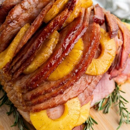 A glazed holiday ham adorned with pineapple slices sits on a wooden board, garnished with sprigs of rosemary.