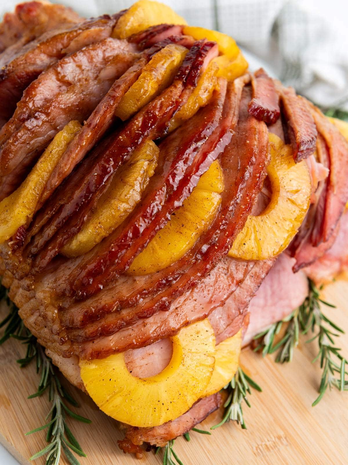 A glazed holiday ham adorned with pineapple slices sits on a wooden board, garnished with sprigs of rosemary.