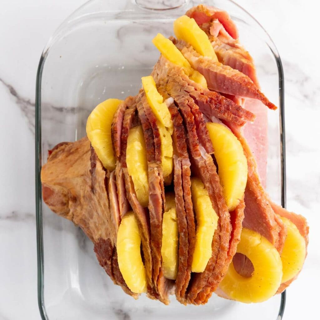 Sliced ham with pineapple rings between the slices, placed in a glass baking dish on a marble countertop.