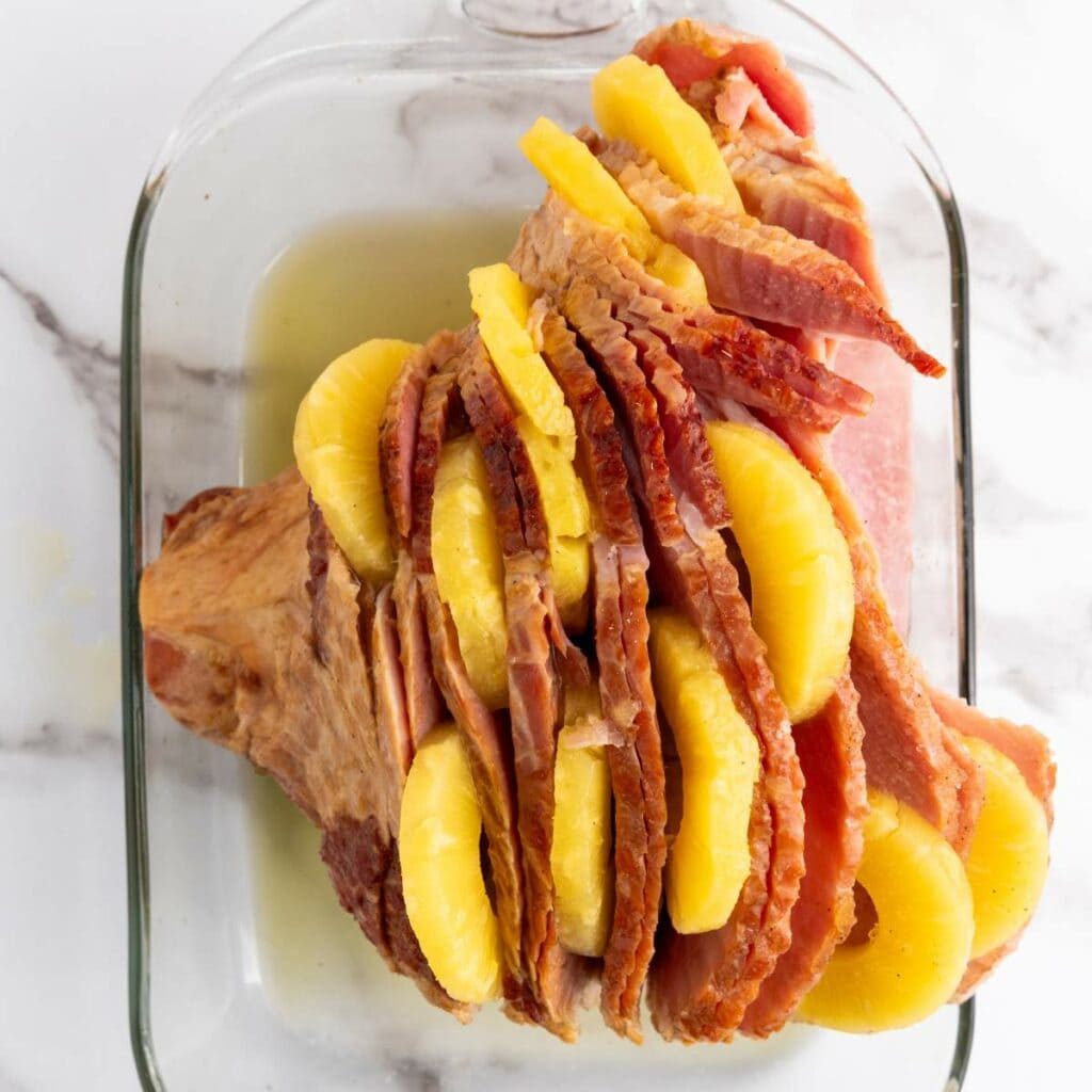 A glazed ham in a glass baking dish, sliced with pineapple rings inserted between the slices.