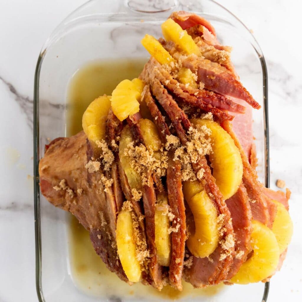 A glazed ham topped with pineapple slices and brown sugar in a glass baking dish.