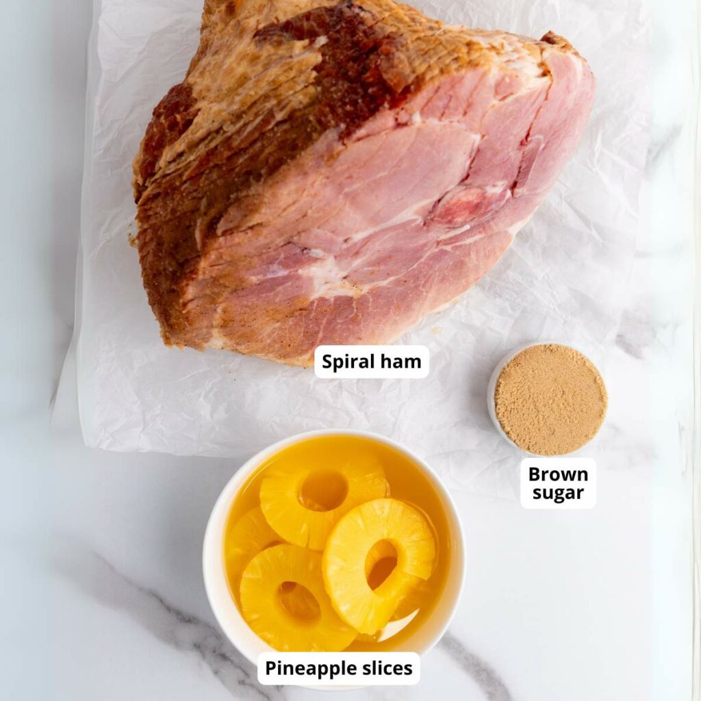 Spiral ham on parchment paper, with a bowl of pineapple slices and a dish of brown sugar.