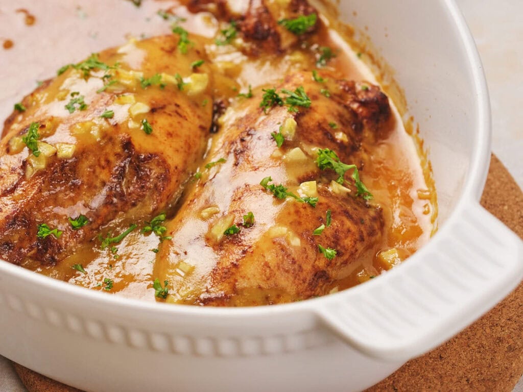 Honey mustard chicken in a white baking dish, covered in a brown sauce and garnished with chopped parsley.