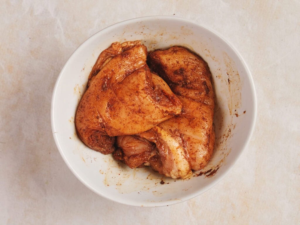 Raw chicken breast marinated with spices in a white bowl on a light countertop.