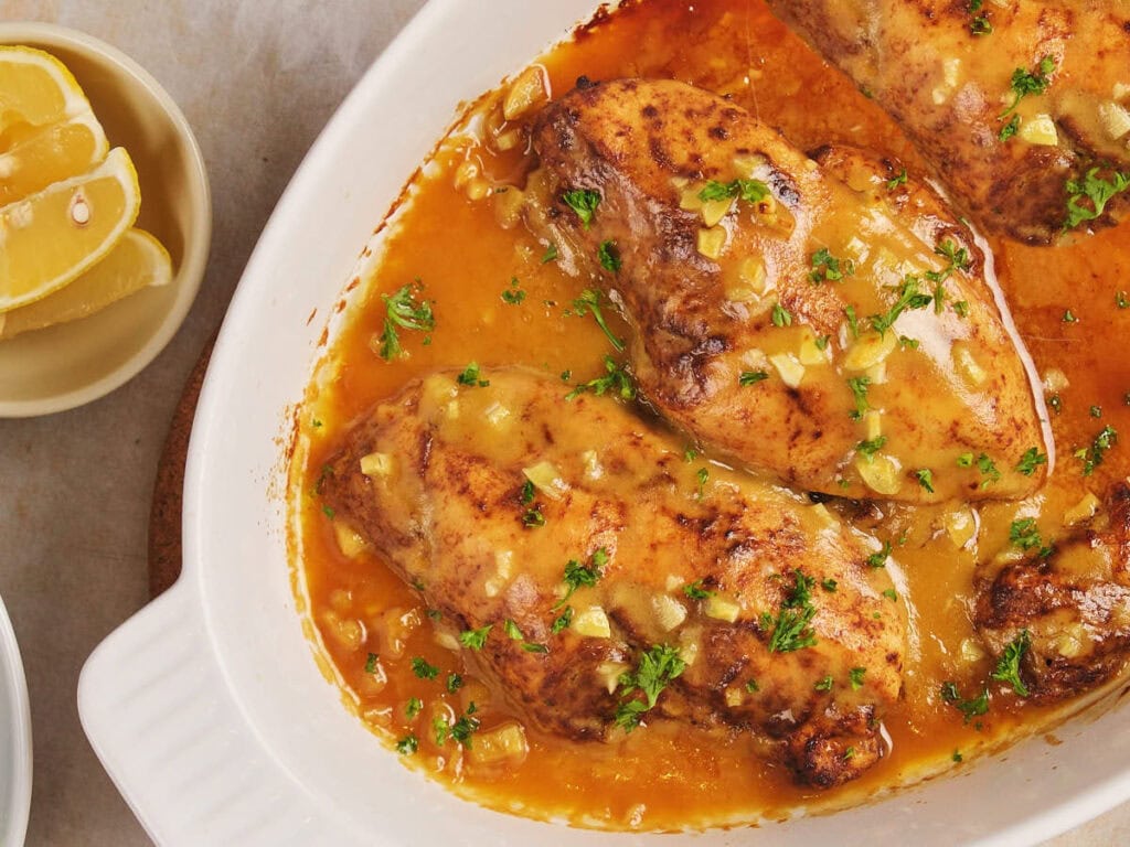 Baked chicken breasts in a lemon garlic sauce, garnished with parsley, in a white dish.