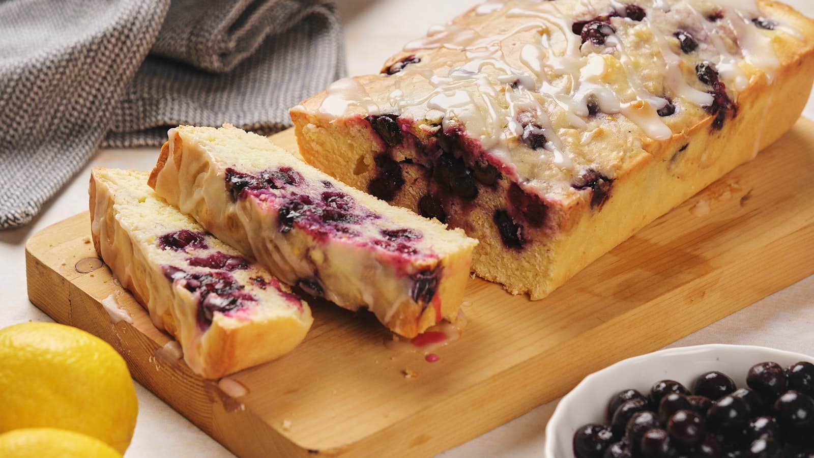 Lemon blueberry pound cake with glaze on a wooden board, partially sliced.