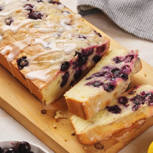 Sliced lemon blueberry pound cake with glaze on a wooden board, surrounded by lemons and blueberries.