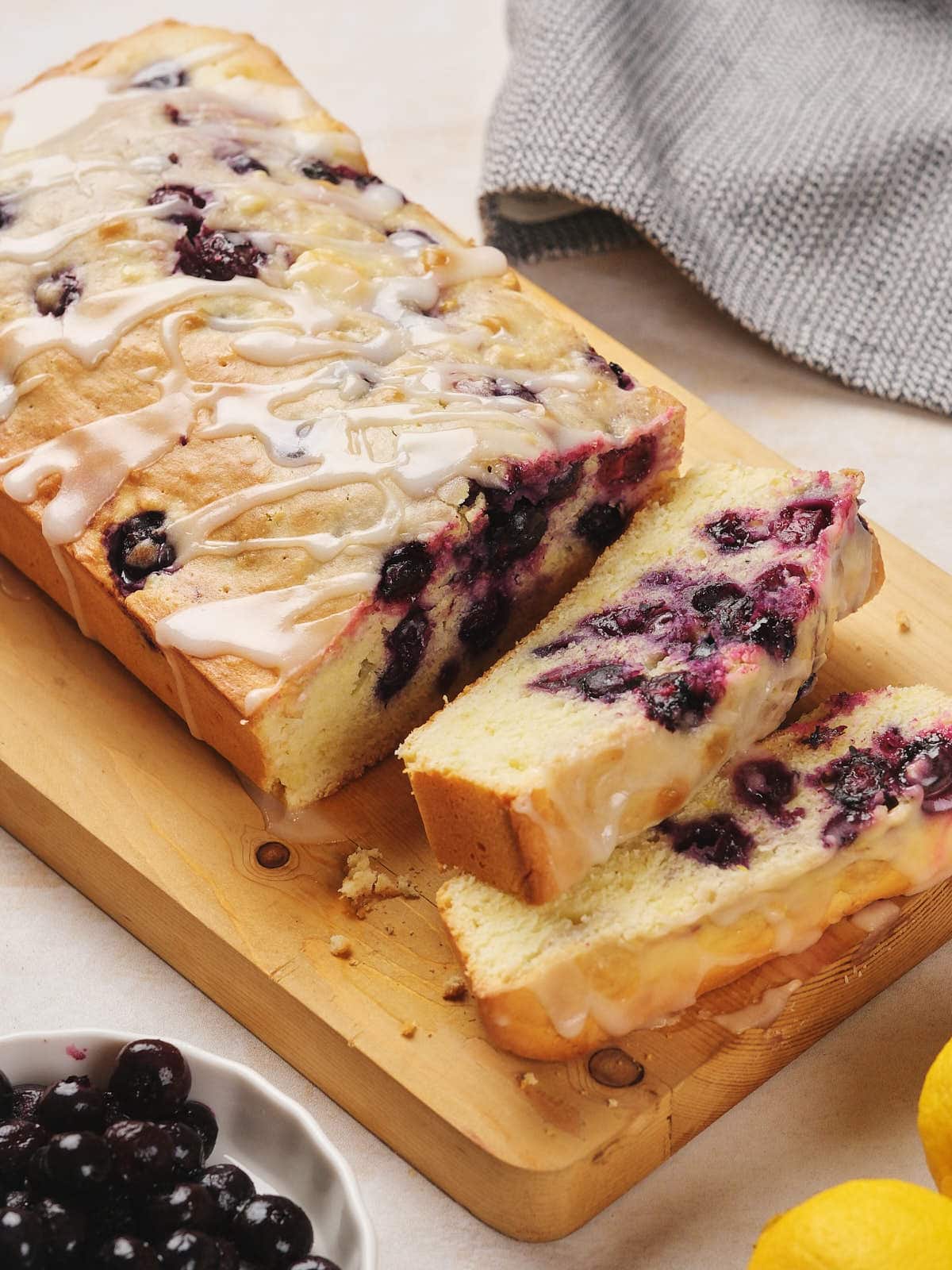 Sliced lemon blueberry pound cake with glaze on a wooden board, surrounded by lemons and blueberries.