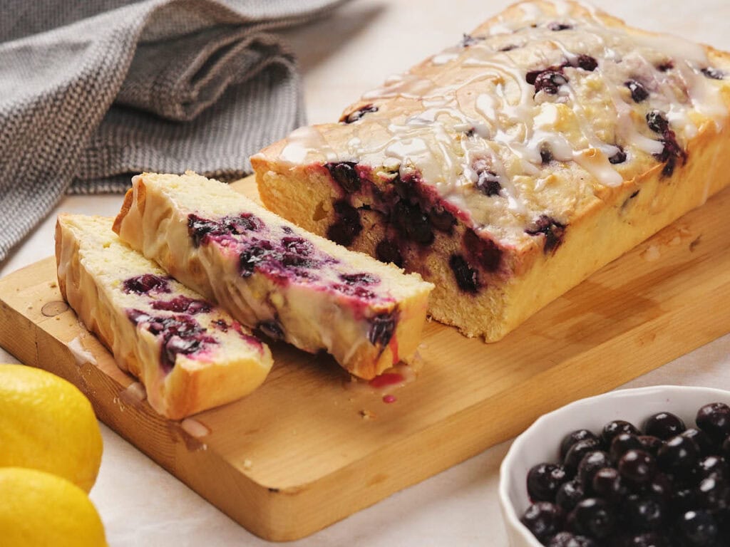 Sliced blueberry loaf cake with glaze on a wooden board. Lemons and a bowl of blueberries are nearby.