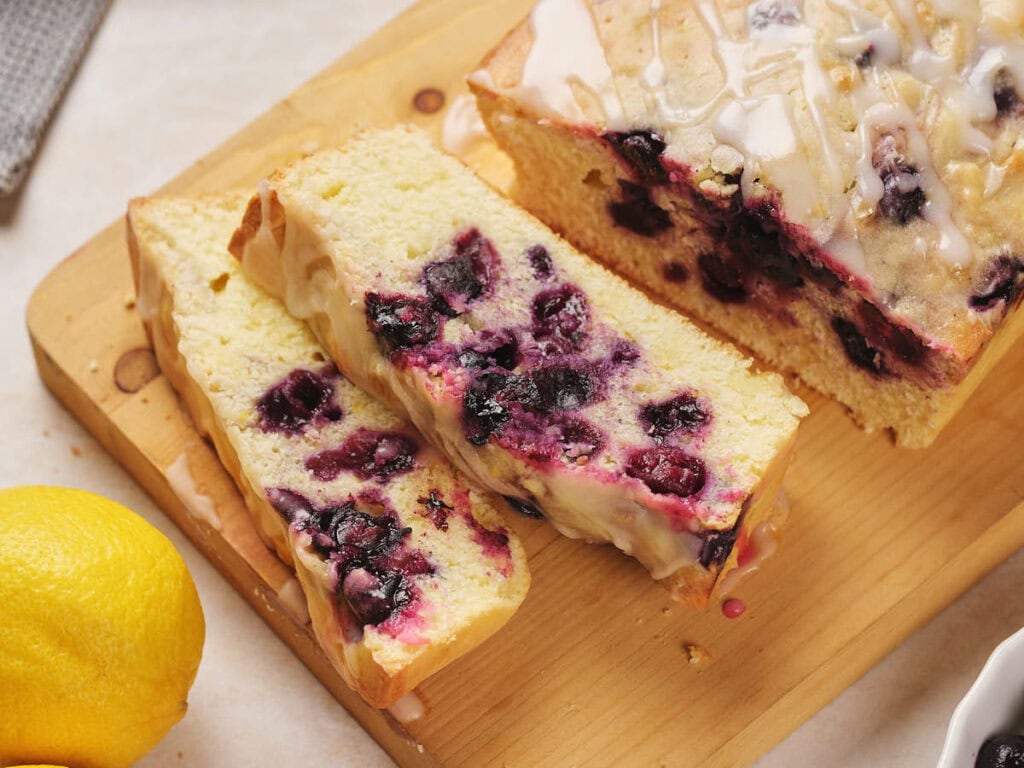 Lemon blueberry pound cake with glaze on a wooden cutting board. Two slices are cut from the loaf.