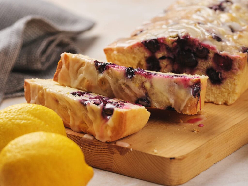Lemon blueberry loaf with glaze on a wooden board, two slices cut. Two whole lemons are nearby.