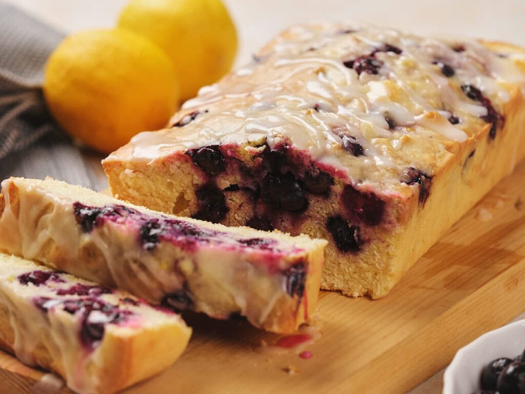 A glazed blueberry lemon loaf cake on a wooden board with two slices cut. Two whole lemons are in the background.
