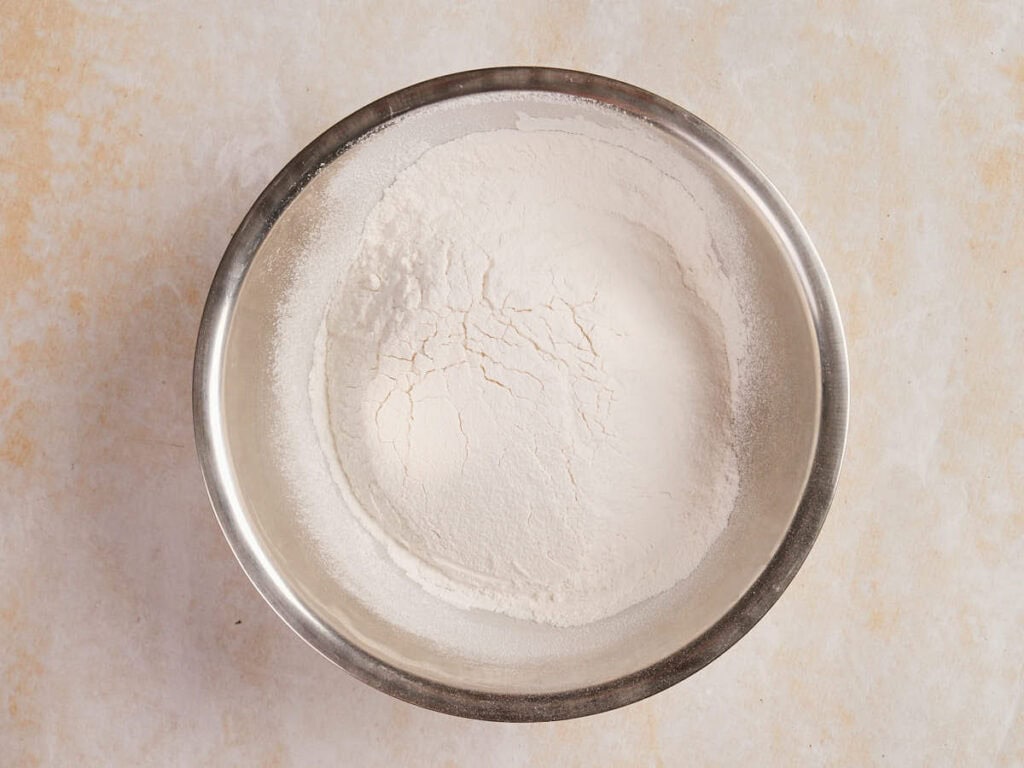 A metal bowl containing a mound of white flour on a light-colored surface.