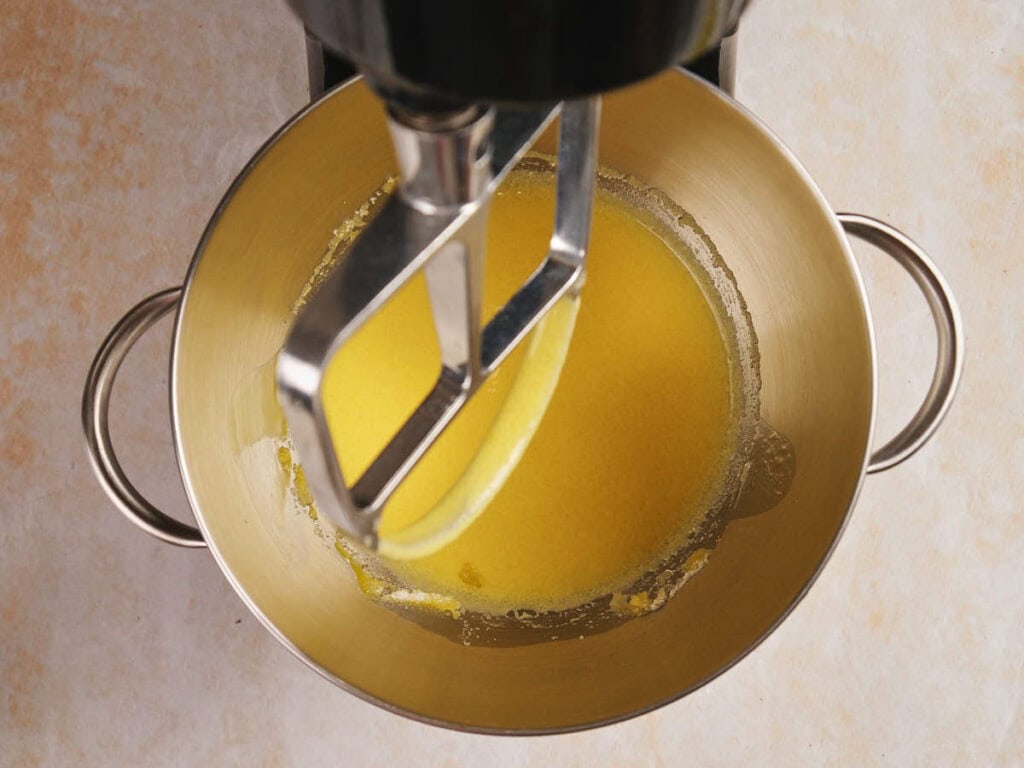 A mixing bowl with handles contains a yellow liquid being mixed by a stand mixer paddle attachment.