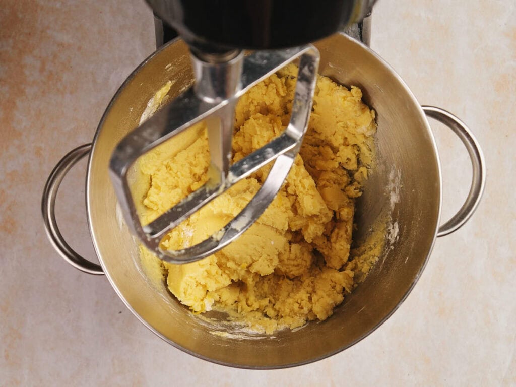 Top view of a mixing bowl with yellow dough being mixed by an electric stand mixer.