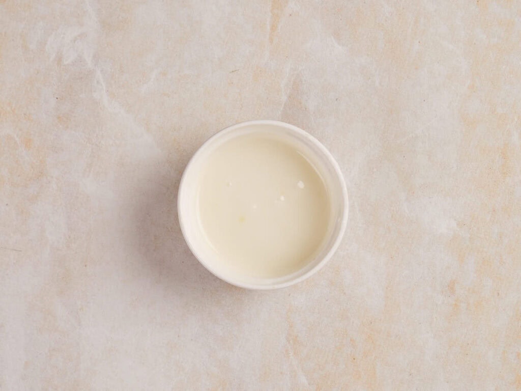 A small white bowl filled with a creamy white liquid on a textured beige surface.