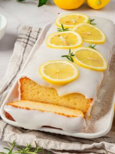 A loaf cake with white icing and lemon slices on top is displayed on a tray. One slice is partially cut in front.