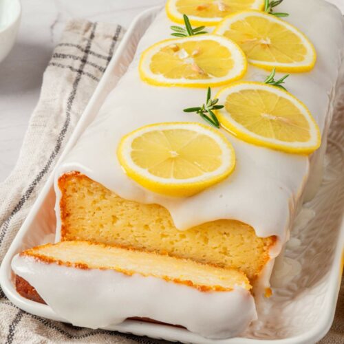 A loaf cake with white icing and lemon slices on top is displayed on a tray. One slice is partially cut in front.