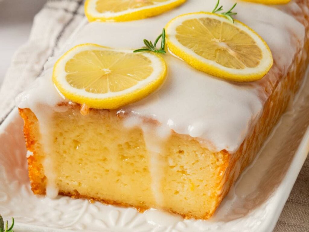 A lemon loaf cake topped with glaze and lemon slices on a decorative white platter.