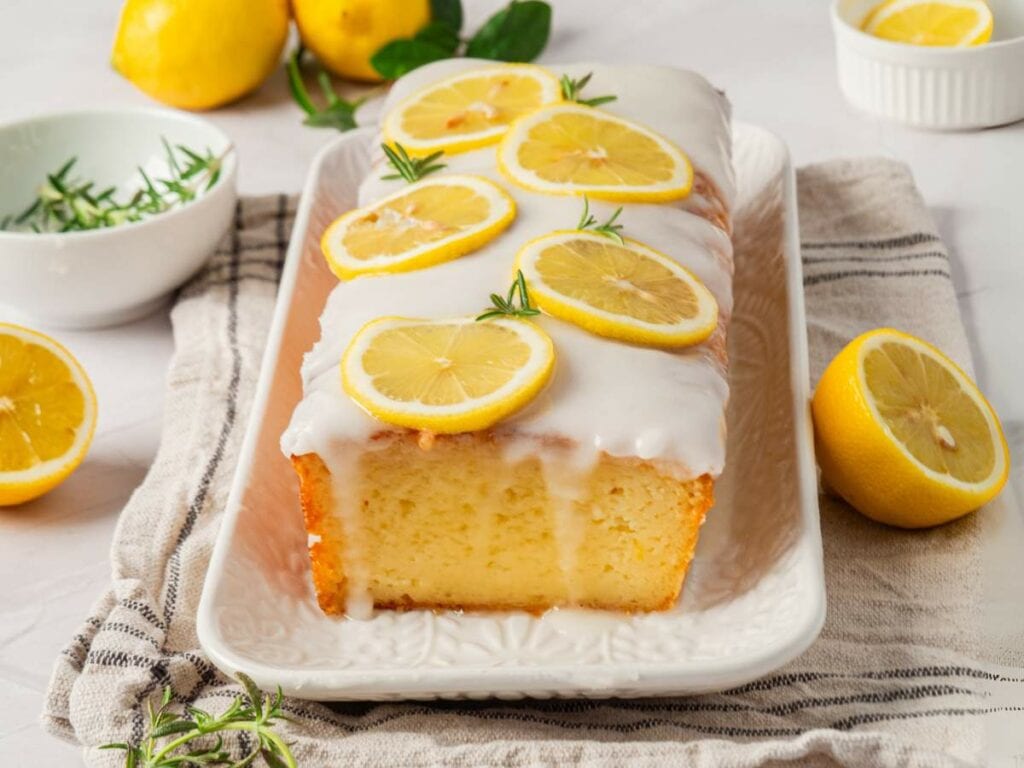 Lemon cake with white glaze, garnished with lemon slices and rosemary, on a white platter.