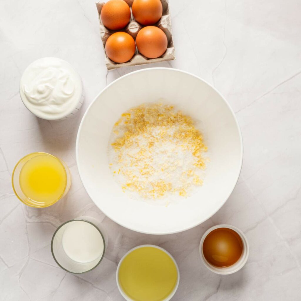 A bowl with dry ingredients surrounded by eggs, yogurt, oil, milk, juice, and vanilla on a light surface.